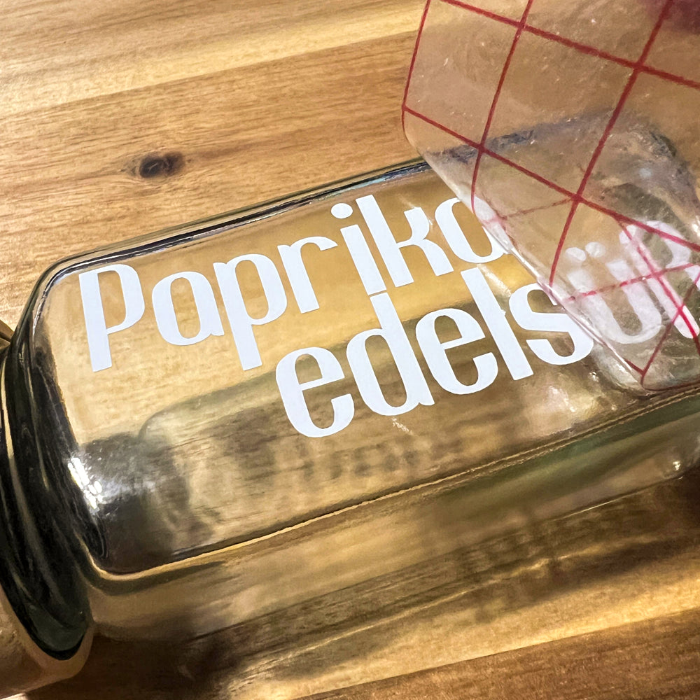 Weißer Vinyl-Name ‚Papriko edelsüß‘ auf einem Glasbehälter, Transferfolie wird gerade abgezogen