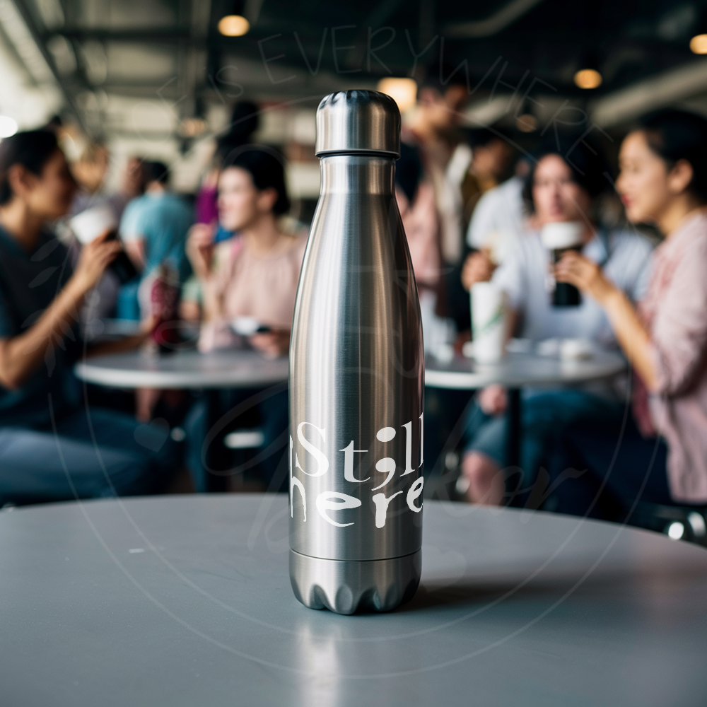 Edelstahl-Trinkflasche mit dem Schriftzug ‚St;ll here‘ auf einem Tisch, Menschen im Hintergrund unscharf