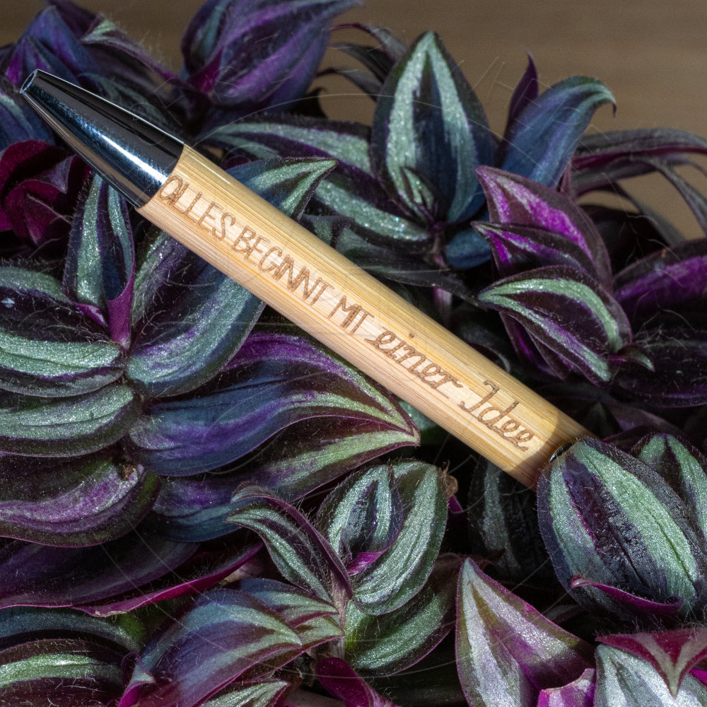 Bambus-Kugelschreiber mit Lasergravur ‚Alles beginnt immer wieder‘, liegend auf violett-grünen Blättern