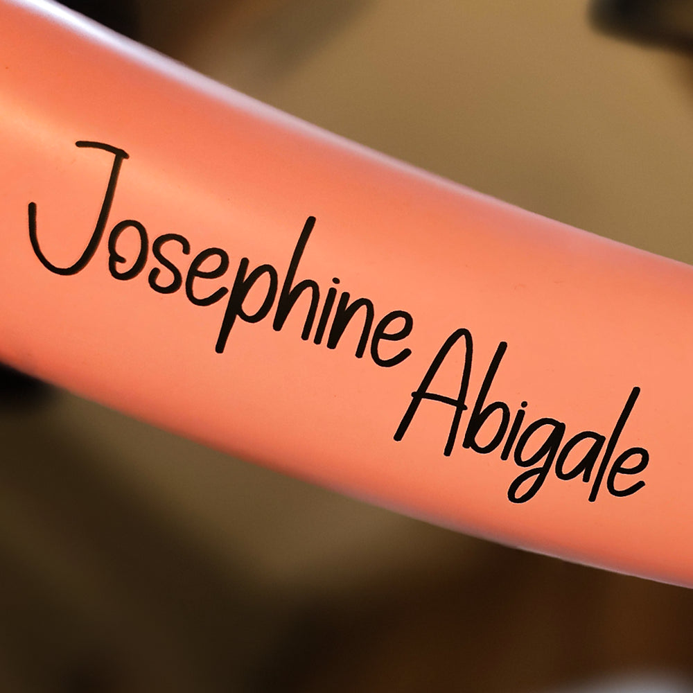 Mittlere Strebe eines Fahrradlaufrads mit schwarzem Vinyl-Namensaufkleber ‚Josephine Abigale‘ auf rosafarbenem Rahmen